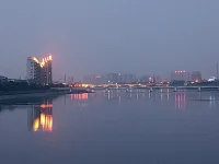 傍晚，我們騎車又來到這里。天色陰暗，整個天空被大霧籠罩，看不到明月與星辰，內心也好似蒙塵。坐了一會兒，已沒有雅興觀賞，往返途中，回頭一望，遠處高樓聳立，樓頂上紅色燈火映在水面上形成一道光束，猶如落日余暉灑在水面上，紅彤彤，金燦燦地，絢爛奪目，水面上依然波光如鏡。驀然心里注入一道光，當你的視覺僅停留在灰色地帶，就難以發現生活的那抹亮色，當你轉念一想，其實好景就在那里，你注意到它，它就給你點亮心情。你看那水，縱然外面煙霧繚繞，我自水波不興，風平浪靜。配圖