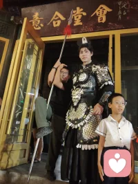 《杨柳青古巷遇戎装赋》
 
暮临津沽杨柳青墟，长街灯华，古肆连楹。余闲步漫游，忽见一戎装少年立巷隅，玄铠鎏纹，长戈倚侧，眉目疏朗，英气凌云。金甲映霓虹，身姿挺拔若青松临风，顾盼之间，俊采飞扬，令人目移心喜。
 
余趋前相与语，言欲留影纪此相逢。少年温雅谦和，闻言欣然颔首，俯仰立坐，皆顺人意，摆姿展态，毫无倦怠，待人以诚，气度温润，初见便觉心头烦扰消解大半。方调镜头之际，一家四口缓步而至，远客夫妻携两稚子，远道来游古镇。夫君执机欲为妻取景，持镜茫然，不知章法。余侧立旁，细教取景光影、身形站位之法，嘱其寻温柔姿态衬佳人风姿。
 
其夫闻言莞尔，回身笑语妻曰：“无妨，可依偎相拥，随心喜乐，不必拘谨。”语罢伸手相挽，妻霎时展颜，笑靥如花，眉眼间羞怯尽散。身旁两童见状，亦拍手嬉闹，绕膝奔走，满是童真烂漫。少顷夫君移步上前，自请立镜前，摹美猴王腾跃之姿，抬手探额，嬉闹无拘，阖家笑语充盈古巷。
 
余立旁静观，心下慨然。此一家四口，夫知惜妻，妻善解意，稚童无忧，彼此体恤成全，无隔阂拘束，方是华夏家庭本真之温情。尘世奔波，世人常困于疏离、苛责，心有郁结，难寻松弛暖意。今古镇灯火、飒爽少年治愈尘俗疲惫，更得此和睦阖家涤荡心绪，恍然知晓治愈本心者，从非远在山海，乃是人间相守的松弛与包容。
 
戎装少年以谦和待人，予过客温柔；寻常夫妇以坦诚相待，予彼此欢愉。烟火古巷，铠甲英姿衬阖家温情，一静一动，皆是人间至善光景。愿九州万户皆如此：夫妻相知不藏拘谨，亲子相伴常存欢颜，家人之间随心抒意，彼此成全，相守和乐，岁岁常有笑语相伴，岁岁皆得温柔治愈。世间万千烦忧，皆可在家人松弛暖意里缓缓消融，人间至幸，莫过于阖家自在，心无芥蒂，喜乐常随。配图