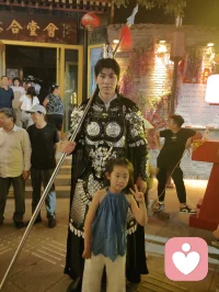 《杨柳青古巷遇戎装赋》
 
暮临津沽杨柳青墟，长街灯华，古肆连楹。余闲步漫游，忽见一戎装少年立巷隅，玄铠鎏纹，长戈倚侧，眉目疏朗，英气凌云。金甲映霓虹，身姿挺拔若青松临风，顾盼之间，俊采飞扬，令人目移心喜。
 
余趋前相与语，言欲留影纪此相逢。少年温雅谦和，闻言欣然颔首，俯仰立坐，皆顺人意，摆姿展态，毫无倦怠，待人以诚，气度温润，初见便觉心头烦扰消解大半。方调镜头之际，一家四口缓步而至，远客夫妻携两稚子，远道来游古镇。夫君执机欲为妻取景，持镜茫然，不知章法。余侧立旁，细教取景光影、身形站位之法，嘱其寻温柔姿态衬佳人风姿。
 
其夫闻言莞尔，回身笑语妻曰：“无妨，可依偎相拥，随心喜乐，不必拘谨。”语罢伸手相挽，妻霎时展颜，笑靥如花，眉眼间羞怯尽散。身旁两童见状，亦拍手嬉闹，绕膝奔走，满是童真烂漫。少顷夫君移步上前，自请立镜前，摹美猴王腾跃之姿，抬手探额，嬉闹无拘，阖家笑语充盈古巷。
 
余立旁静观，心下慨然。此一家四口，夫知惜妻，妻善解意，稚童无忧，彼此体恤成全，无隔阂拘束，方是华夏家庭本真之温情。尘世奔波，世人常困于疏离、苛责，心有郁结，难寻松弛暖意。今古镇灯火、飒爽少年治愈尘俗疲惫，更得此和睦阖家涤荡心绪，恍然知晓治愈本心者，从非远在山海，乃是人间相守的松弛与包容。
 
戎装少年以谦和待人，予过客温柔；寻常夫妇以坦诚相待，予彼此欢愉。烟火古巷，铠甲英姿衬阖家温情，一静一动，皆是人间至善光景。愿九州万户皆如此：夫妻相知不藏拘谨，亲子相伴常存欢颜，家人之间随心抒意，彼此成全，相守和乐，岁岁常有笑语相伴，岁岁皆得温柔治愈。世间万千烦忧，皆可在家人松弛暖意里缓缓消融，人间至幸，莫过于阖家自在，心无芥蒂，喜乐常随。配图