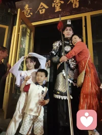 《杨柳青古巷遇戎装赋》
 
暮临津沽杨柳青墟，长街灯华，古肆连楹。余闲步漫游，忽见一戎装少年立巷隅，玄铠鎏纹，长戈倚侧，眉目疏朗，英气凌云。金甲映霓虹，身姿挺拔若青松临风，顾盼之间，俊采飞扬，令人目移心喜。
 
余趋前相与语，言欲留影纪此相逢。少年温雅谦和，闻言欣然颔首，俯仰立坐，皆顺人意，摆姿展态，毫无倦怠，待人以诚，气度温润，初见便觉心头烦扰消解大半。方调镜头之际，一家四口缓步而至，远客夫妻携两稚子，远道来游古镇。夫君执机欲为妻取景，持镜茫然，不知章法。余侧立旁，细教取景光影、身形站位之法，嘱其寻温柔姿态衬佳人风姿。
 
其夫闻言莞尔，回身笑语妻曰：“无妨，可依偎相拥，随心喜乐，不必拘谨。”语罢伸手相挽，妻霎时展颜，笑靥如花，眉眼间羞怯尽散。身旁两童见状，亦拍手嬉闹，绕膝奔走，满是童真烂漫。少顷夫君移步上前，自请立镜前，摹美猴王腾跃之姿，抬手探额，嬉闹无拘，阖家笑语充盈古巷。
 
余立旁静观，心下慨然。此一家四口，夫知惜妻，妻善解意，稚童无忧，彼此体恤成全，无隔阂拘束，方是华夏家庭本真之温情。尘世奔波，世人常困于疏离、苛责，心有郁结，难寻松弛暖意。今古镇灯火、飒爽少年治愈尘俗疲惫，更得此和睦阖家涤荡心绪，恍然知晓治愈本心者，从非远在山海，乃是人间相守的松弛与包容。
 
戎装少年以谦和待人，予过客温柔；寻常夫妇以坦诚相待，予彼此欢愉。烟火古巷，铠甲英姿衬阖家温情，一静一动，皆是人间至善光景。愿九州万户皆如此：夫妻相知不藏拘谨，亲子相伴常存欢颜，家人之间随心抒意，彼此成全，相守和乐，岁岁常有笑语相伴，岁岁皆得温柔治愈。世间万千烦忧，皆可在家人松弛暖意里缓缓消融，人间至幸，莫过于阖家自在，心无芥蒂，喜乐常随。配图