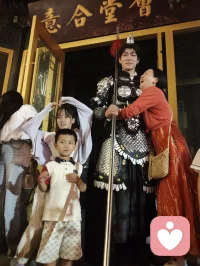 《杨柳青古巷遇戎装赋》
 
暮临津沽杨柳青墟，长街灯华，古肆连楹。余闲步漫游，忽见一戎装少年立巷隅，玄铠鎏纹，长戈倚侧，眉目疏朗，英气凌云。金甲映霓虹，身姿挺拔若青松临风，顾盼之间，俊采飞扬，令人目移心喜。
 
余趋前相与语，言欲留影纪此相逢。少年温雅谦和，闻言欣然颔首，俯仰立坐，皆顺人意，摆姿展态，毫无倦怠，待人以诚，气度温润，初见便觉心头烦扰消解大半。方调镜头之际，一家四口缓步而至，远客夫妻携两稚子，远道来游古镇。夫君执机欲为妻取景，持镜茫然，不知章法。余侧立旁，细教取景光影、身形站位之法，嘱其寻温柔姿态衬佳人风姿。
 
其夫闻言莞尔，回身笑语妻曰：“无妨，可依偎相拥，随心喜乐，不必拘谨。”语罢伸手相挽，妻霎时展颜，笑靥如花，眉眼间羞怯尽散。身旁两童见状，亦拍手嬉闹，绕膝奔走，满是童真烂漫。少顷夫君移步上前，自请立镜前，摹美猴王腾跃之姿，抬手探额，嬉闹无拘，阖家笑语充盈古巷。
 
余立旁静观，心下慨然。此一家四口，夫知惜妻，妻善解意，稚童无忧，彼此体恤成全，无隔阂拘束，方是华夏家庭本真之温情。尘世奔波，世人常困于疏离、苛责，心有郁结，难寻松弛暖意。今古镇灯火、飒爽少年治愈尘俗疲惫，更得此和睦阖家涤荡心绪，恍然知晓治愈本心者，从非远在山海，乃是人间相守的松弛与包容。
 
戎装少年以谦和待人，予过客温柔；寻常夫妇以坦诚相待，予彼此欢愉。烟火古巷，铠甲英姿衬阖家温情，一静一动，皆是人间至善光景。愿九州万户皆如此：夫妻相知不藏拘谨，亲子相伴常存欢颜，家人之间随心抒意，彼此成全，相守和乐，岁岁常有笑语相伴，岁岁皆得温柔治愈。世间万千烦忧，皆可在家人松弛暖意里缓缓消融，人间至幸，莫过于阖家自在，心无芥蒂，喜乐常随。配图